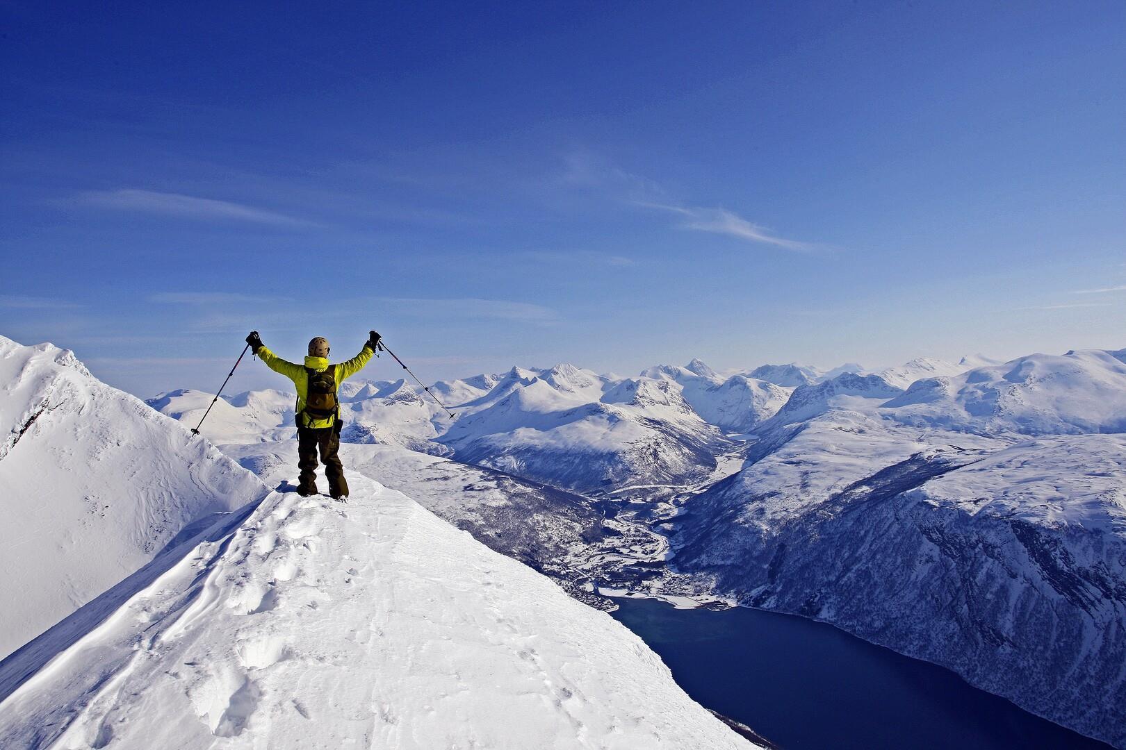 Skiløper løfter stavene til værs på toppen av en kant med snø. I det fjerne fjellene innover Sverige, i forgrunnen nede til høyre skimtes Beisfjord. Bildet er tatt en klar vinterdag med sol.