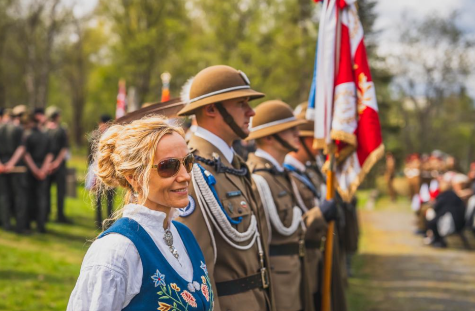 Bilde av Lena Hope som står ved siden av soldater på markering av 28. mai. De står ute, på rekke og rad i ført uniform. De holder flagg. Lena ser i kamera og smiler. - Klikk for stort bilde