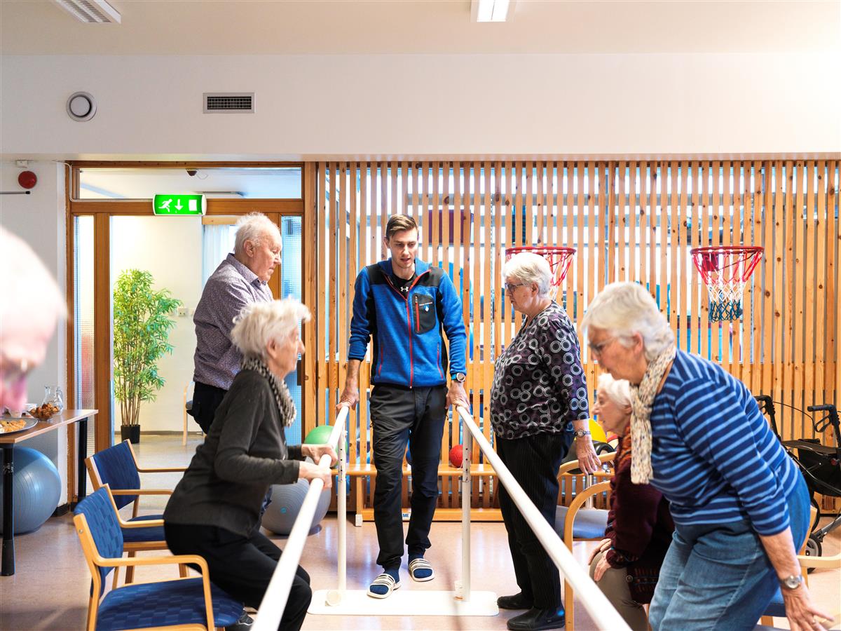 Bilde fra fallforebyggende trening. Bildet viser instruktør i midten, og gruppe med seniorer som står på hver sin side av et rekkverk og trener på enkle øvelser. - Klikk for stort bilde