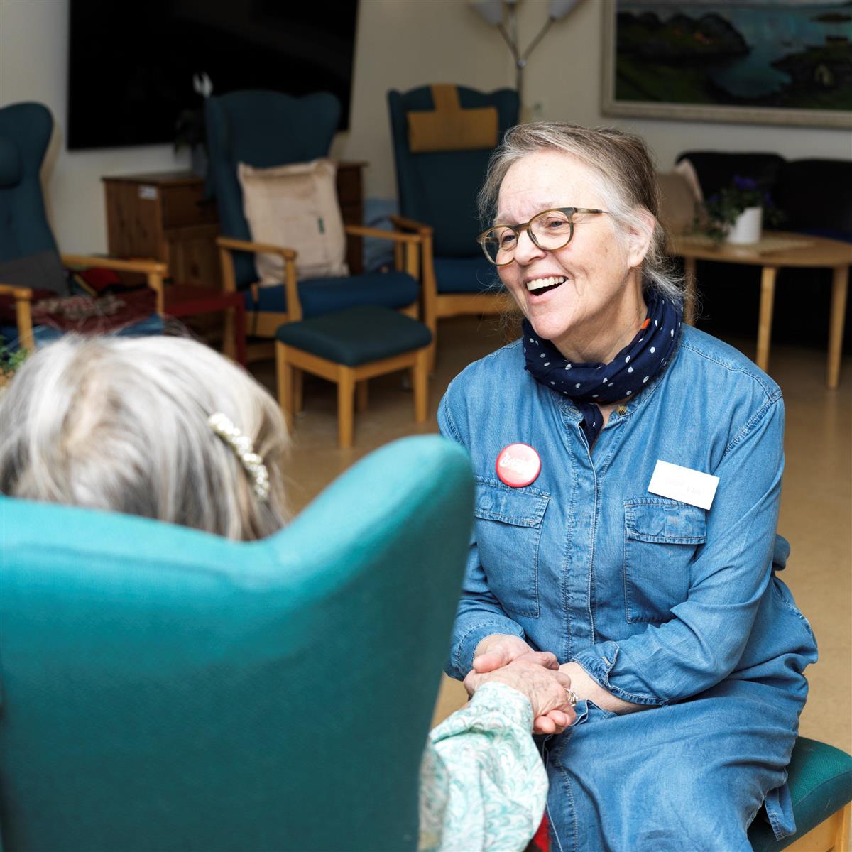 Ansatt på Furumoen holder hånden til en bruker av dagsenteret. Bruker sitter i en stol med ryggen til. I bakgrunnen lenestoler på dagsenteret. - Klikk for stort bilde